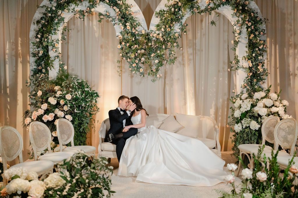 Bride and groom on a sofa, kissing under floral heart arches at a wedding ceremony.