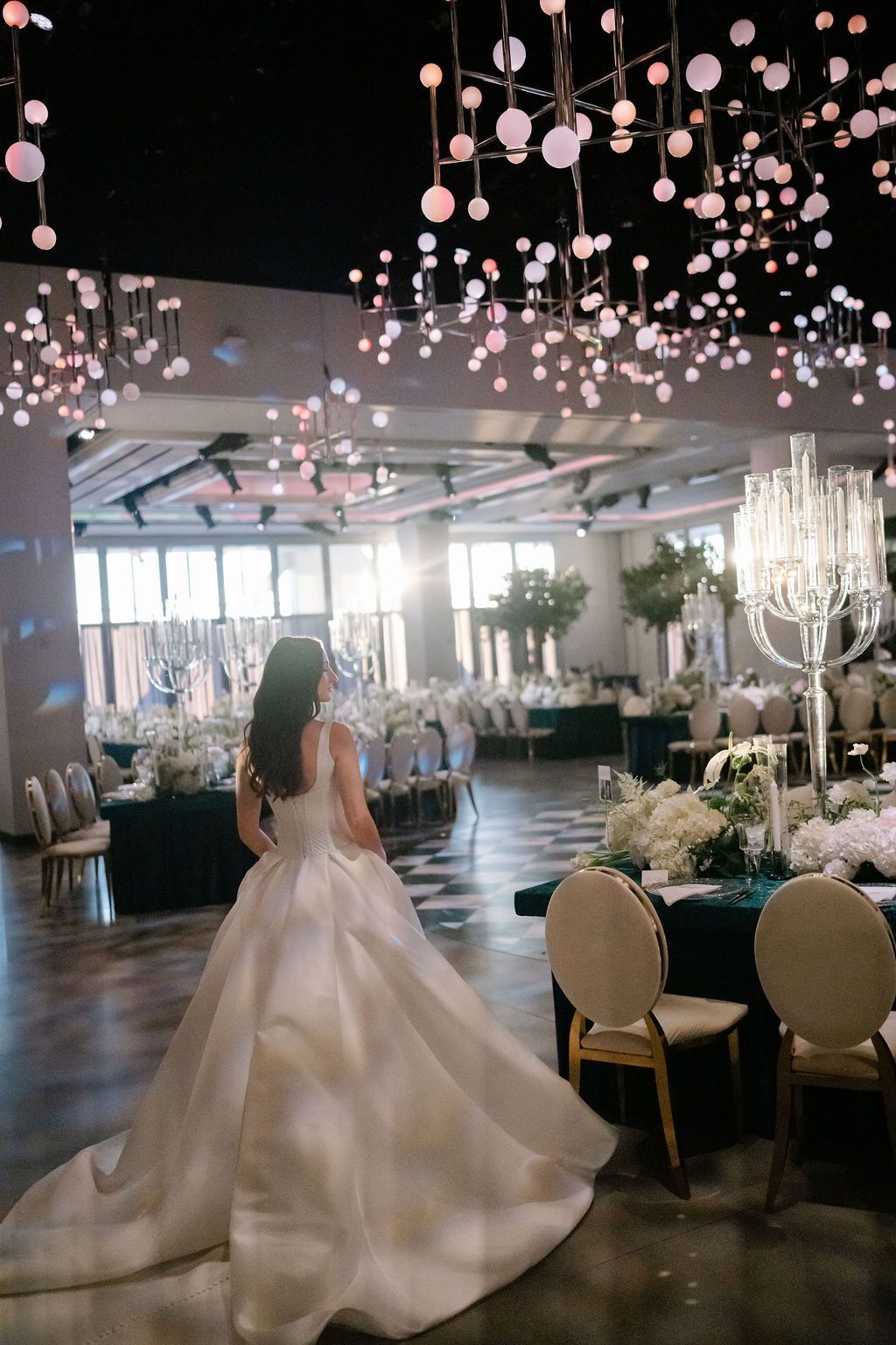 Bride in wedding dress, ballroom with tables, chandelier, and modern light fixtures.
