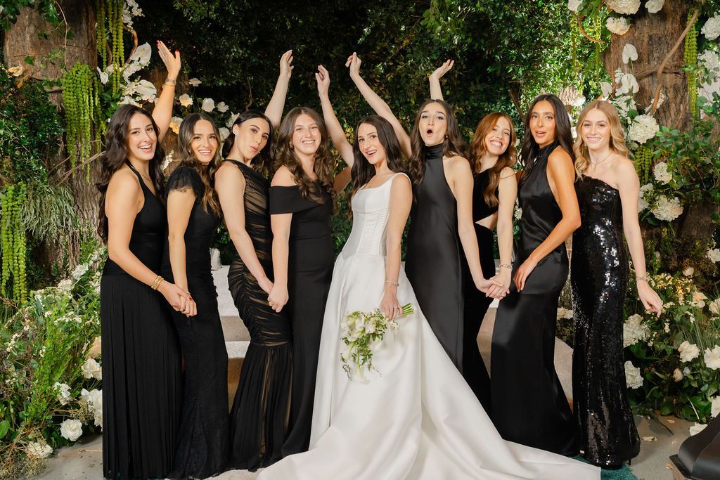 Bride with bridesmaids in black gowns, arms raised, celebrating. Lush floral backdrop.