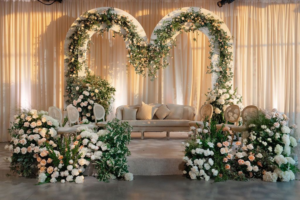 Wedding stage with floral arches, draped backdrop, sofa, and abundant white flowers.