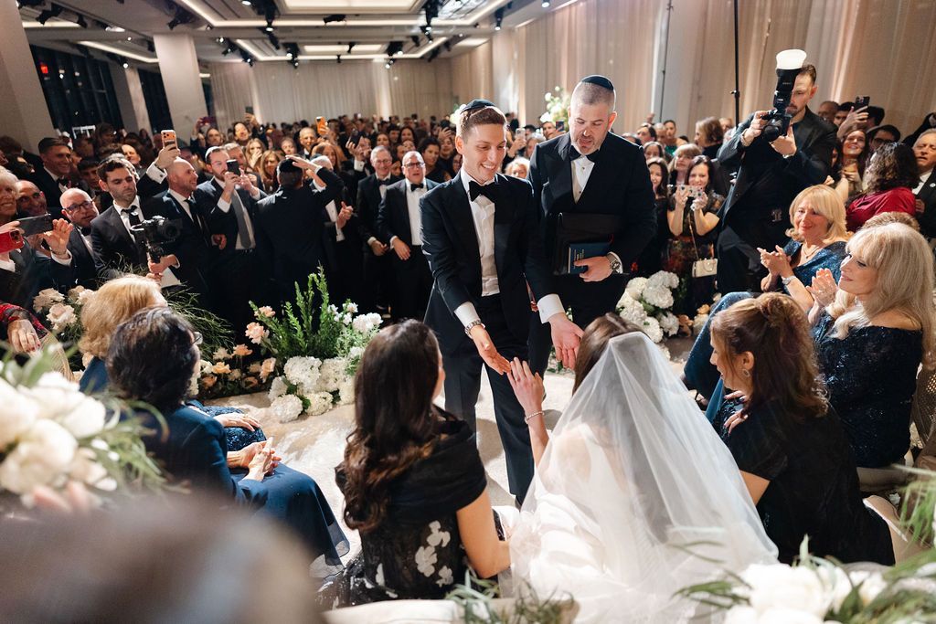 Wedding ceremony: Two men in tuxedos with a bride in a veil surrounded by guests, indoors.