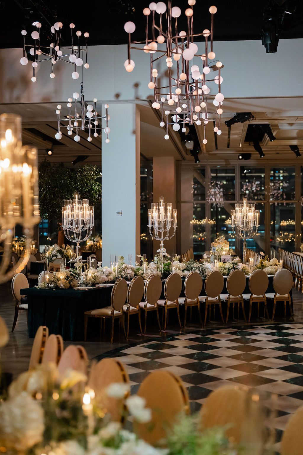 Elegant wedding reception: long table set with flowers, candles, and gold chairs, under modern chandeliers.