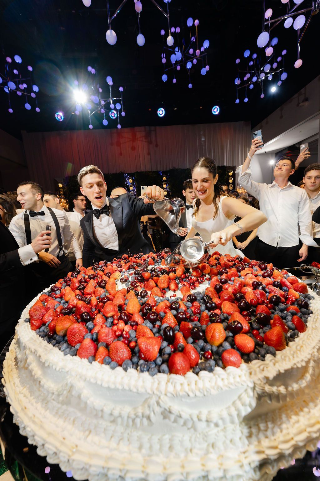 Bride and groom cutting a large cake covered in berries, guests celebrating.