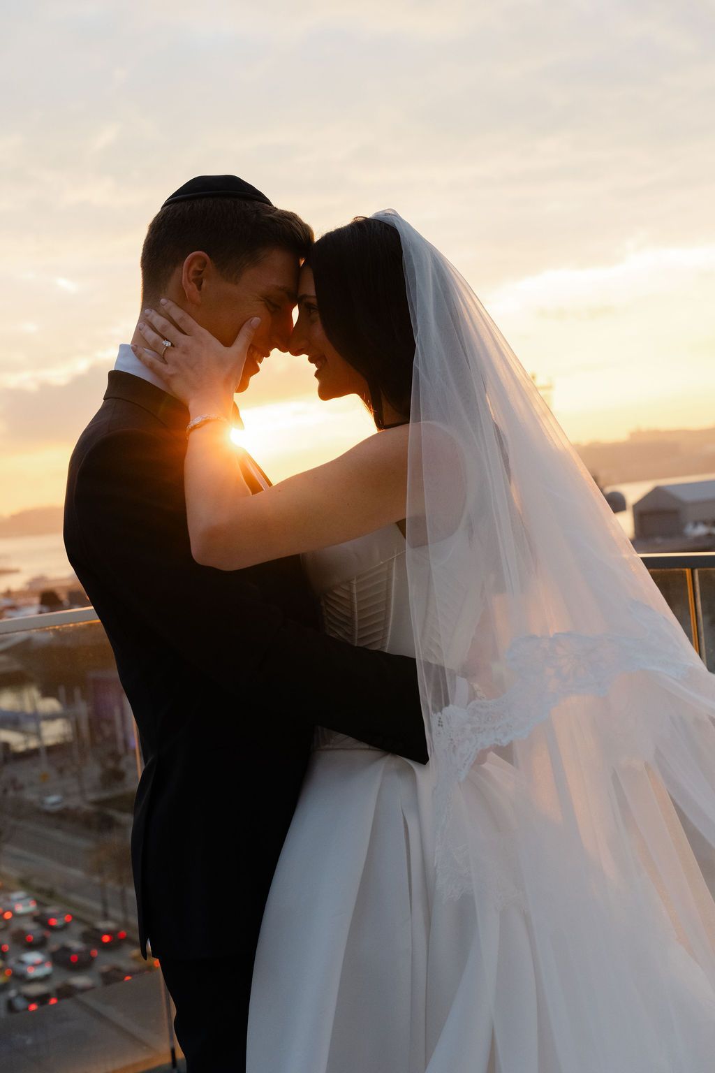 Couple embraces at sunset on rooftop, bride in white dress, groom in suit.