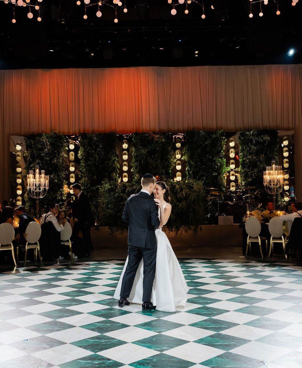 Couple dancing at a wedding reception; checkered dance floor, greenery wall backdrop, and guests at tables.
