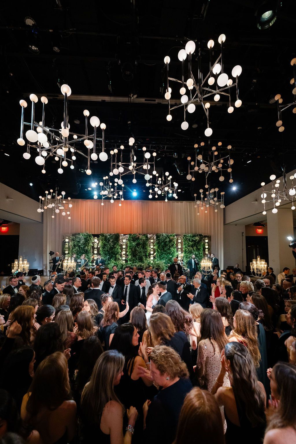 Large event space with many people gathered under decorative lighting. Green backdrop.