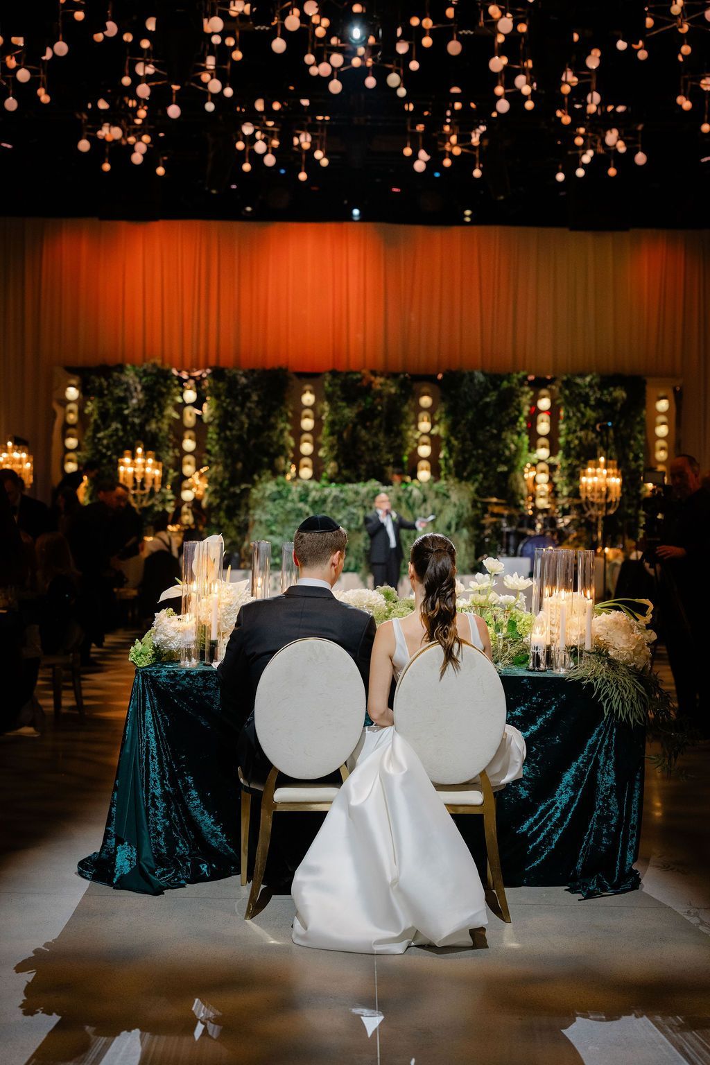 Wedding reception: couple seated, back to camera, at decorated table, greenery backdrop, sparkling lights.