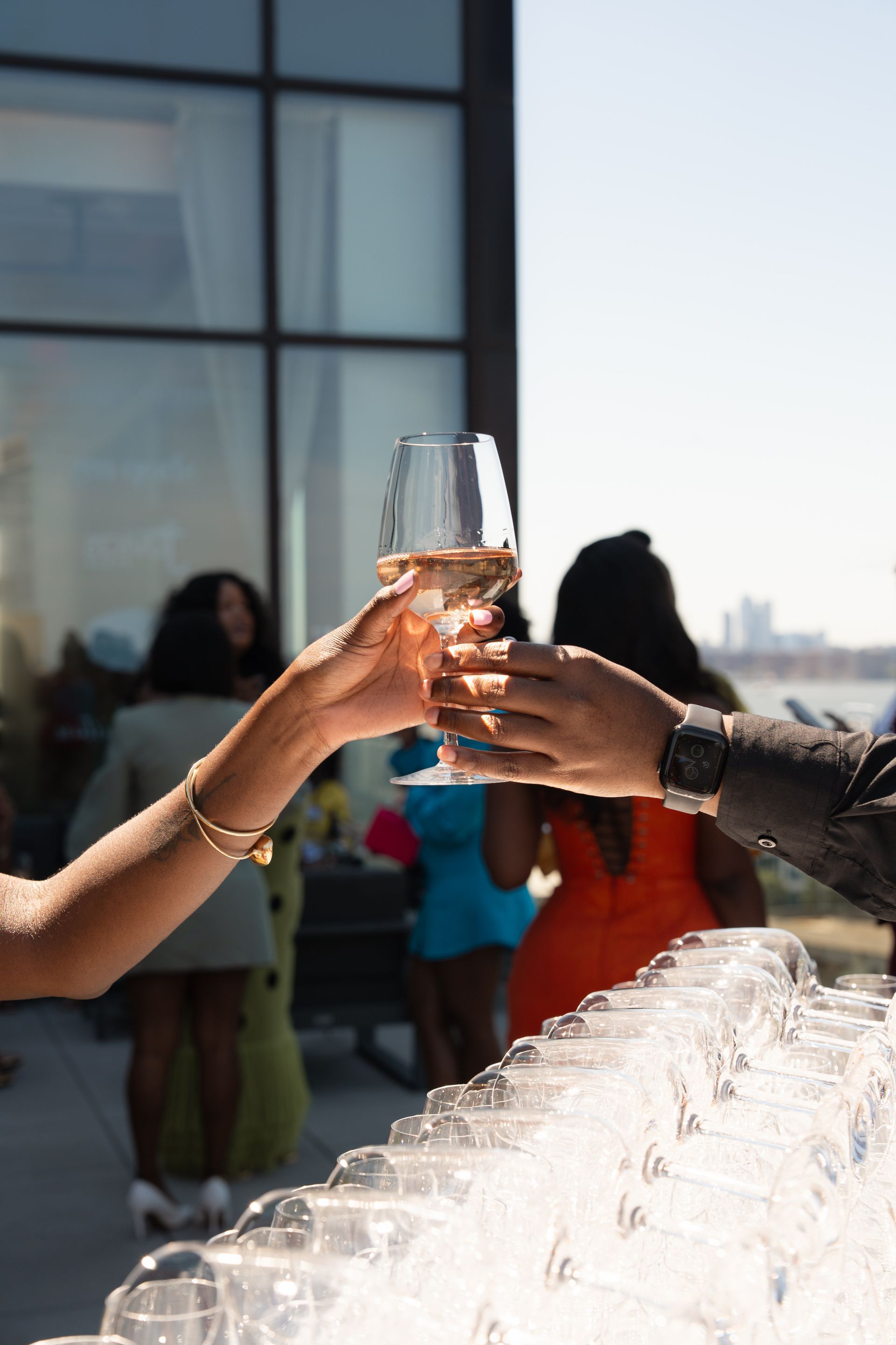 Two people toasting with wine glasses, with other people in the background at an outdoor gathering.