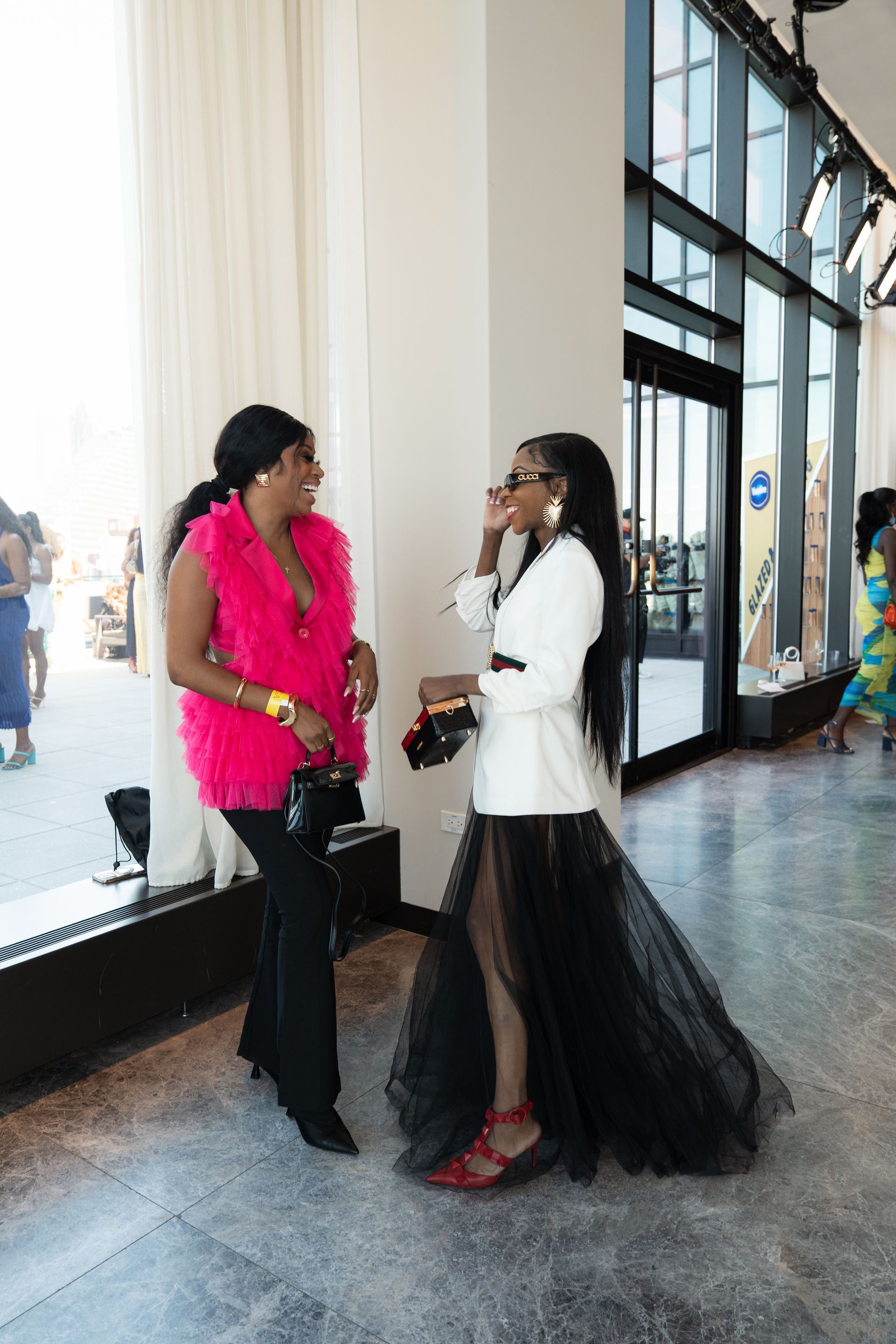 Two women conversing near a window, one in pink fur and black pants, the other in a white jacket and black skirt.