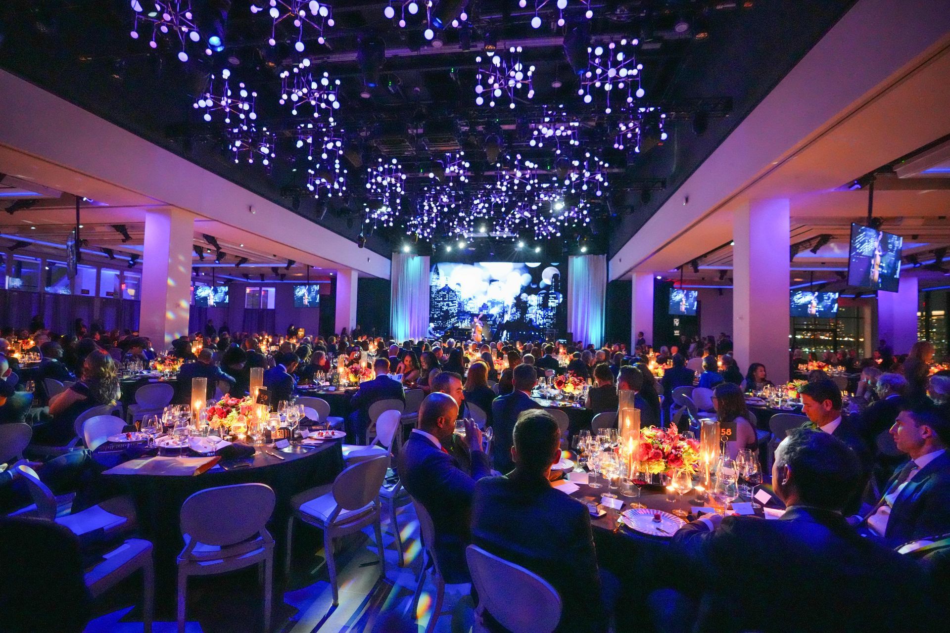 Large banquet hall with many people seated at round tables. Purple lighting and a stage are visible.
