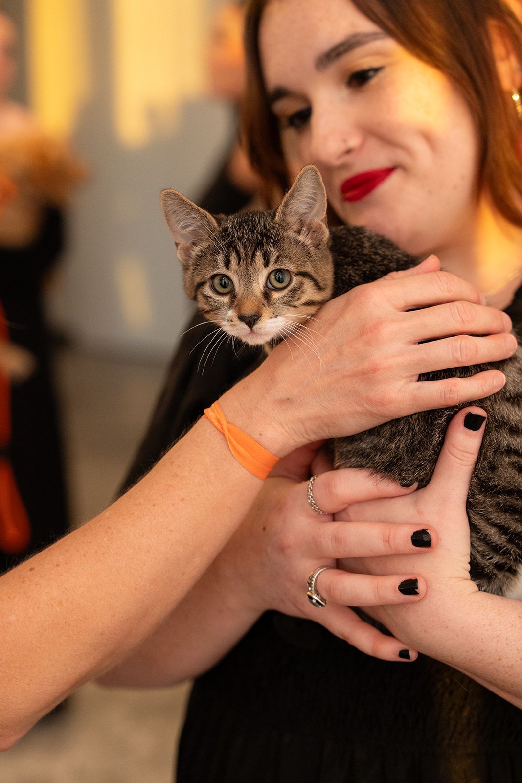 Woman holding a tabby kitten, both looking at the camera. Woman wears black dress, red lipstick, and orange bracelet.