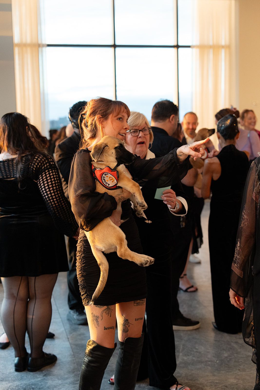 Woman in black dress holds small tan dog, pointing. Another person, also pointing, stands nearby. Indoor gathering.