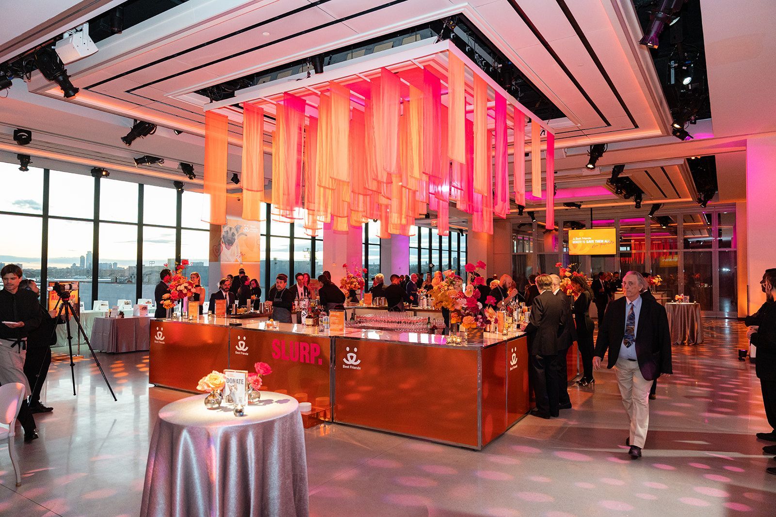 Event reception with bar, floral arrangements, and pink lighting. People gather, windows in background.