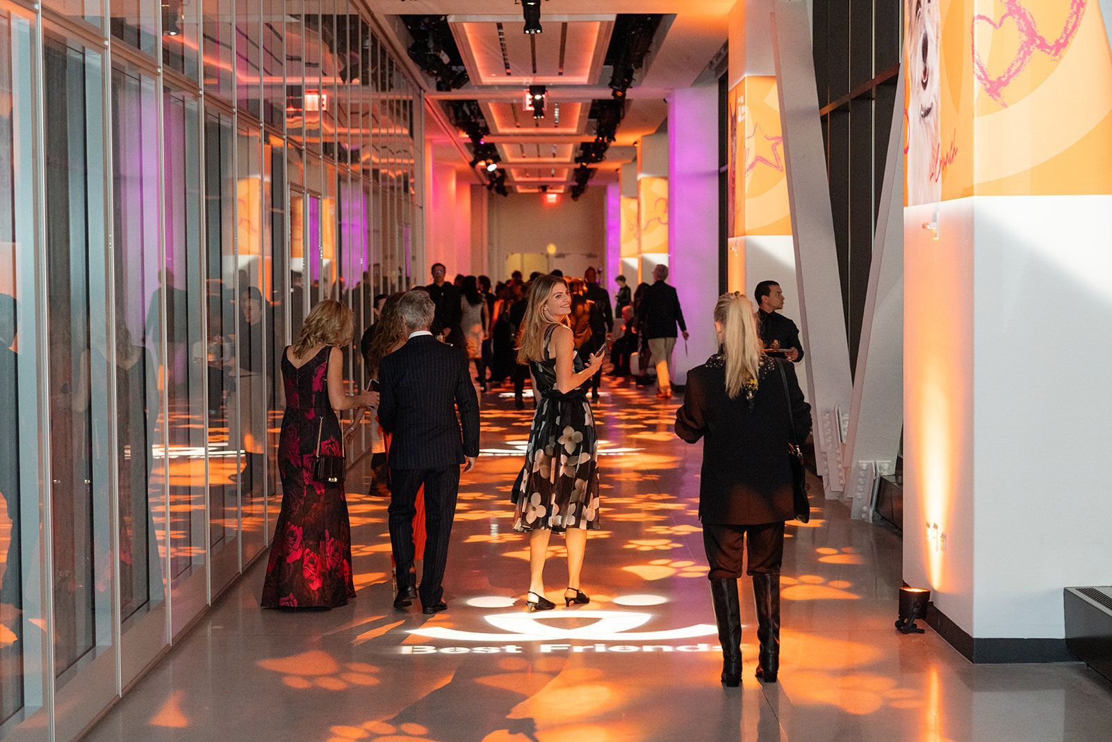 People in formal attire walk down a brightly lit hallway with projected designs.