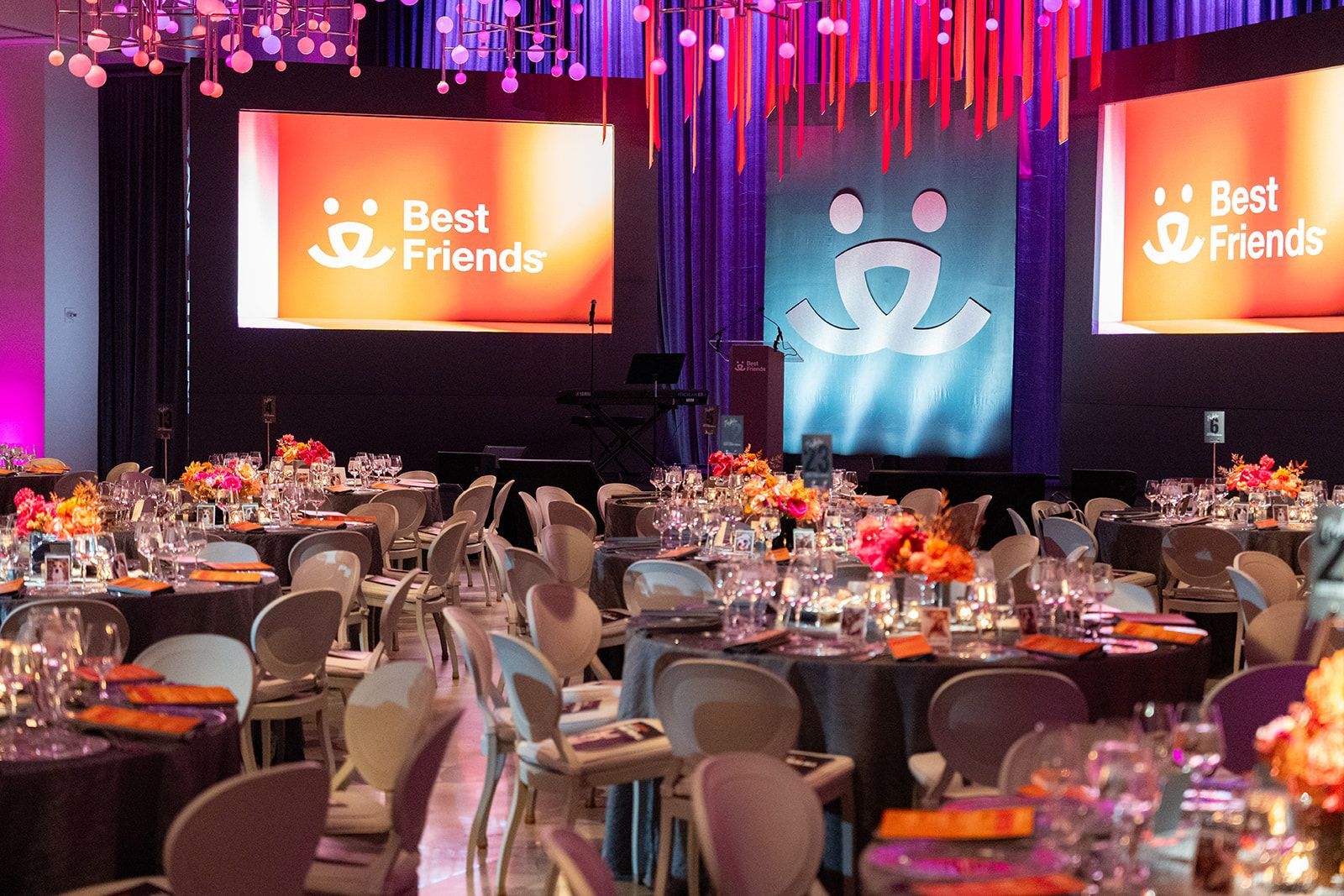 Formal dining event. Tables set with flowers, Best Friends logo on screens, purple and orange decor.