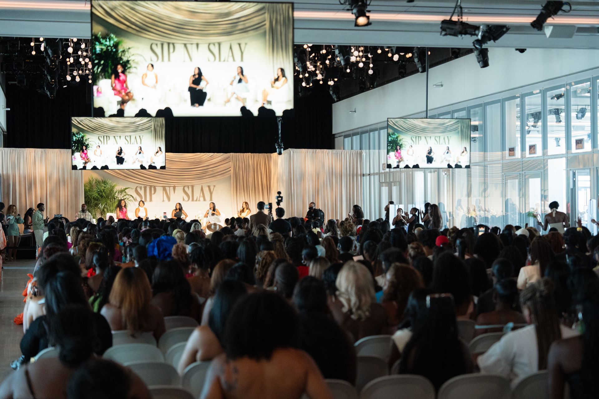Event "Sip 'N' Slay": Women sit in chairs, watching a panel on stage; large screens show the panel.