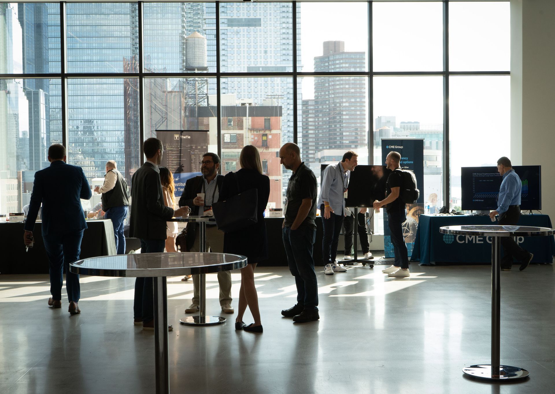 A group of people are standing around tables in a room with a large window.