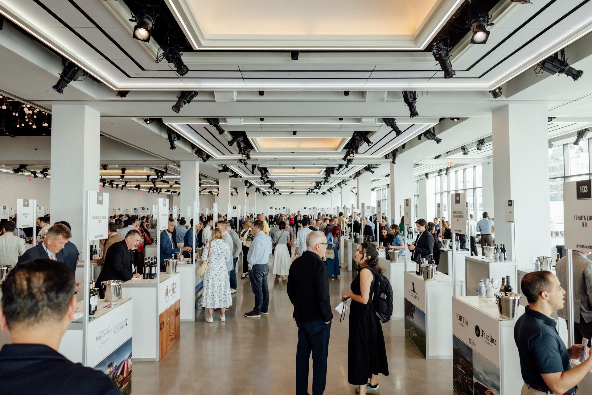 People at a wine tasting event. Large room with tasting tables, bottles, and many attendees. Bright lighting.