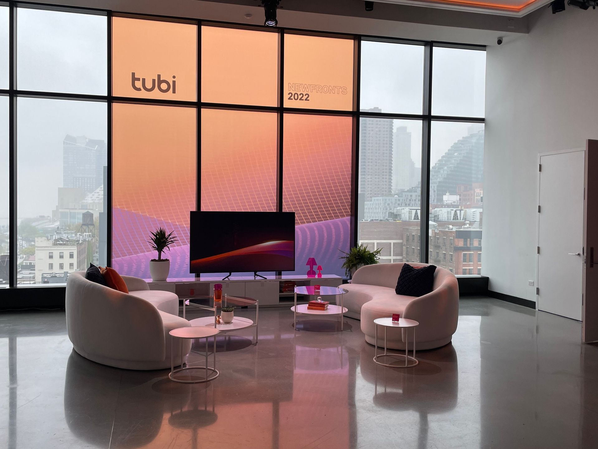 A modern lounge with curved white sofas, small tables, a TV, and a large screen displaying the Tubi logo, with city views.