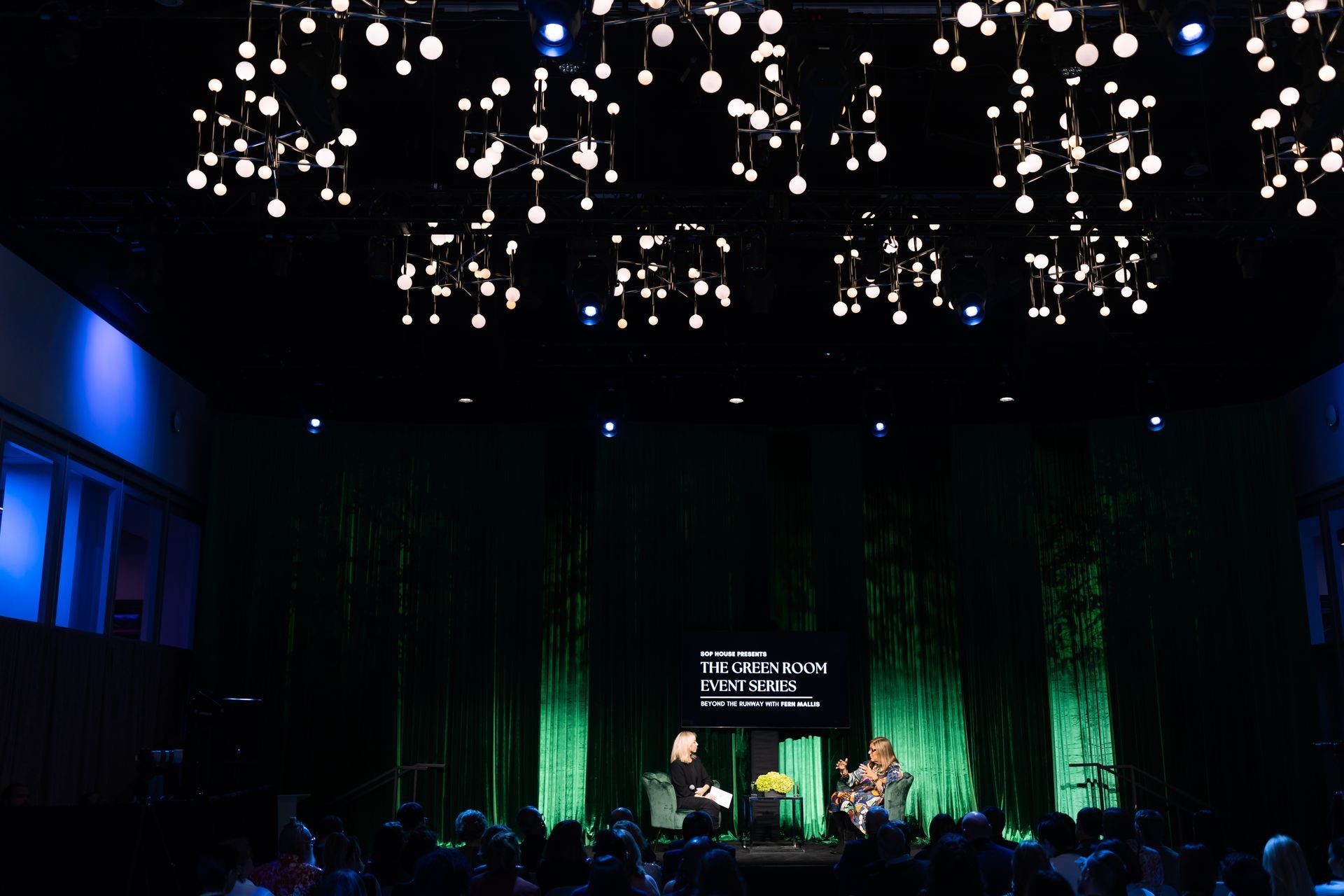 A group of people are sitting in front of a stage with a green curtain.