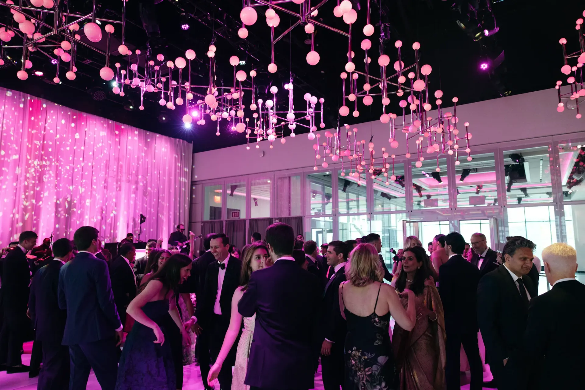 A group of people are dancing in a large room with pink lights hanging from the ceiling.