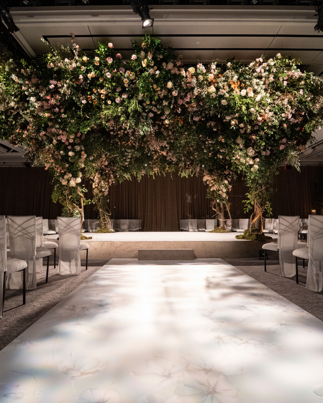 A wedding ceremony is taking place in a large room with flowers hanging from the ceiling.