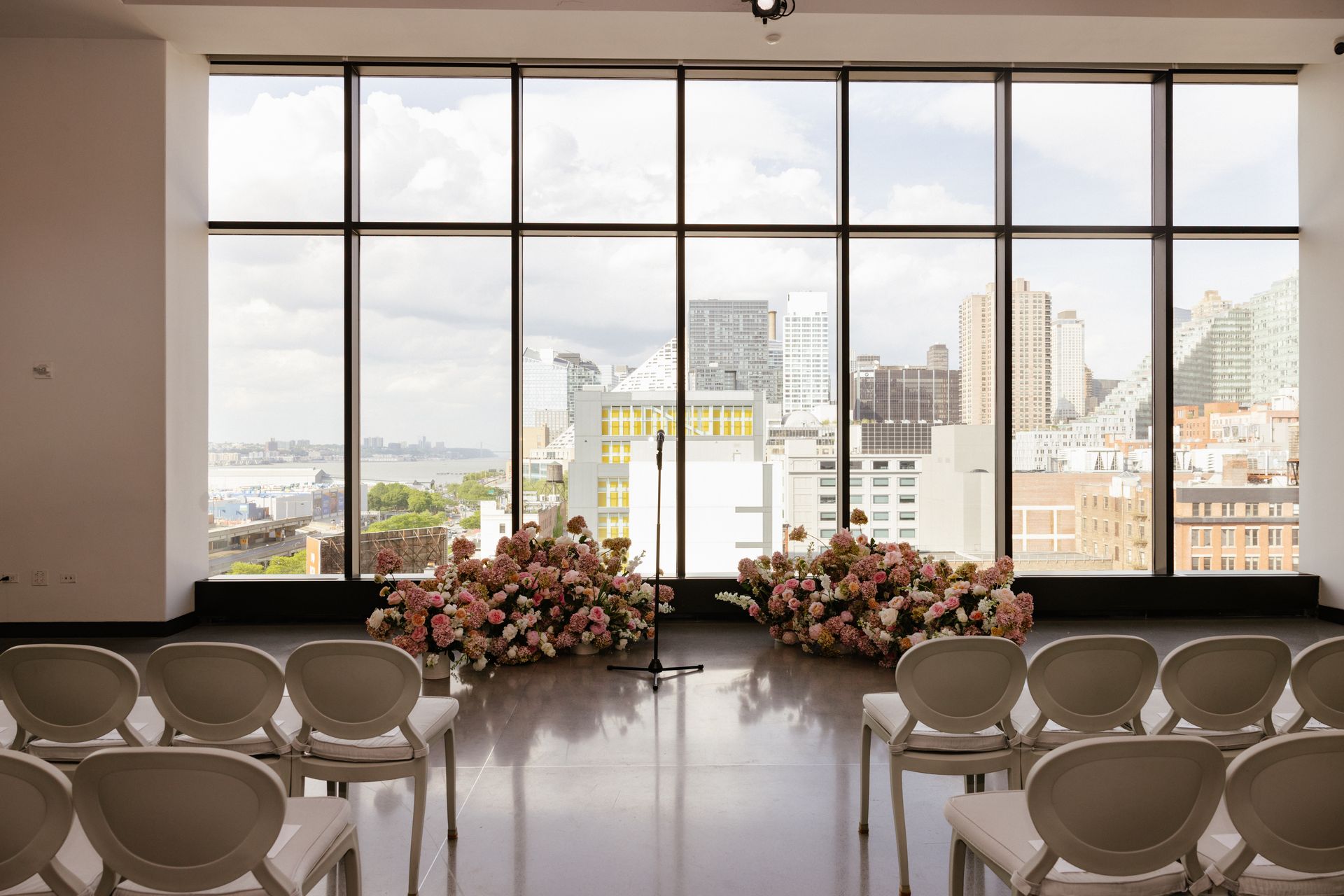 Wedding ceremony setup in a room with a cityscape view, floral arrangements, and white chairs.