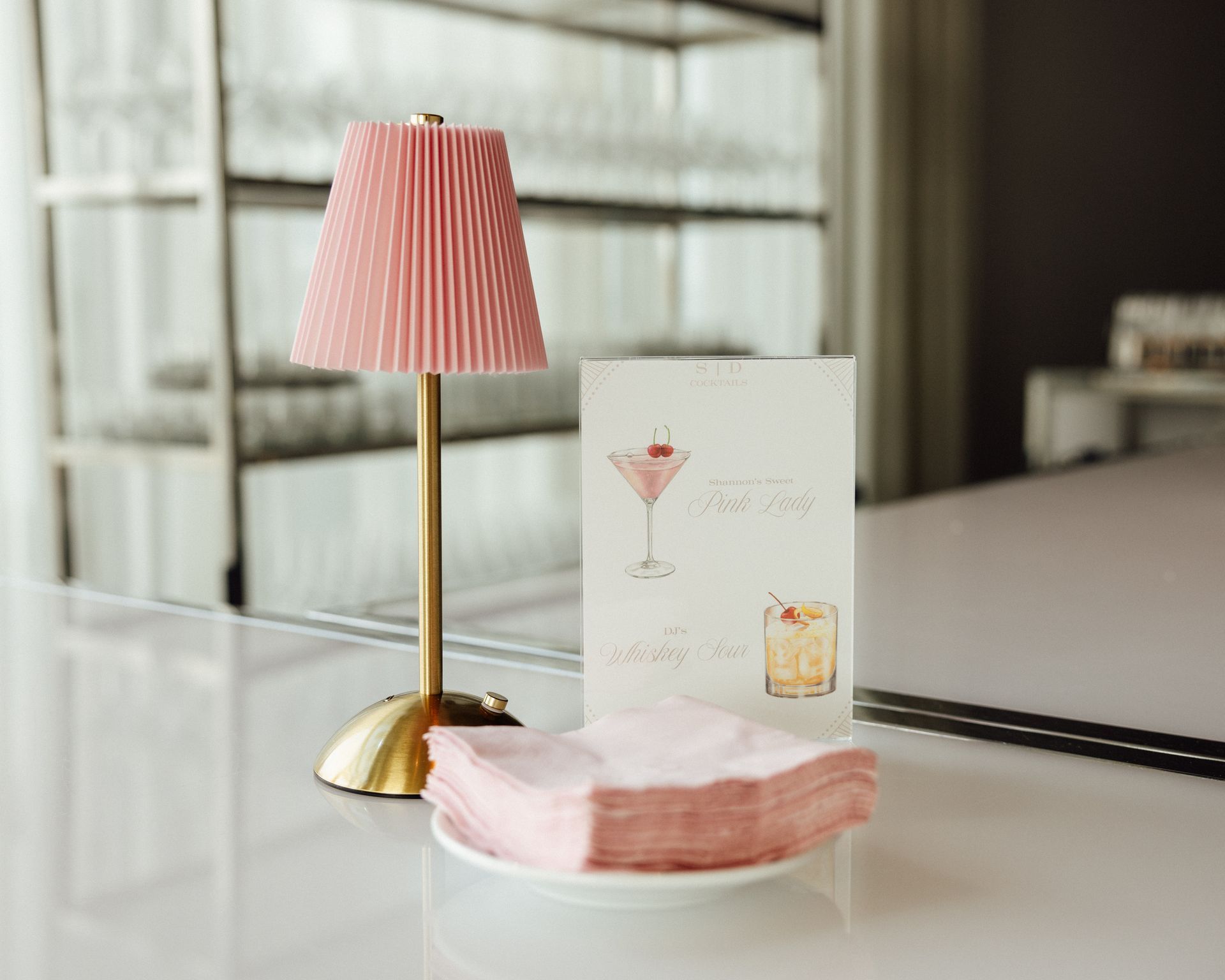 A pink lamp, cocktail menu, and pink napkins on a bar. Background has glassware on shelves.