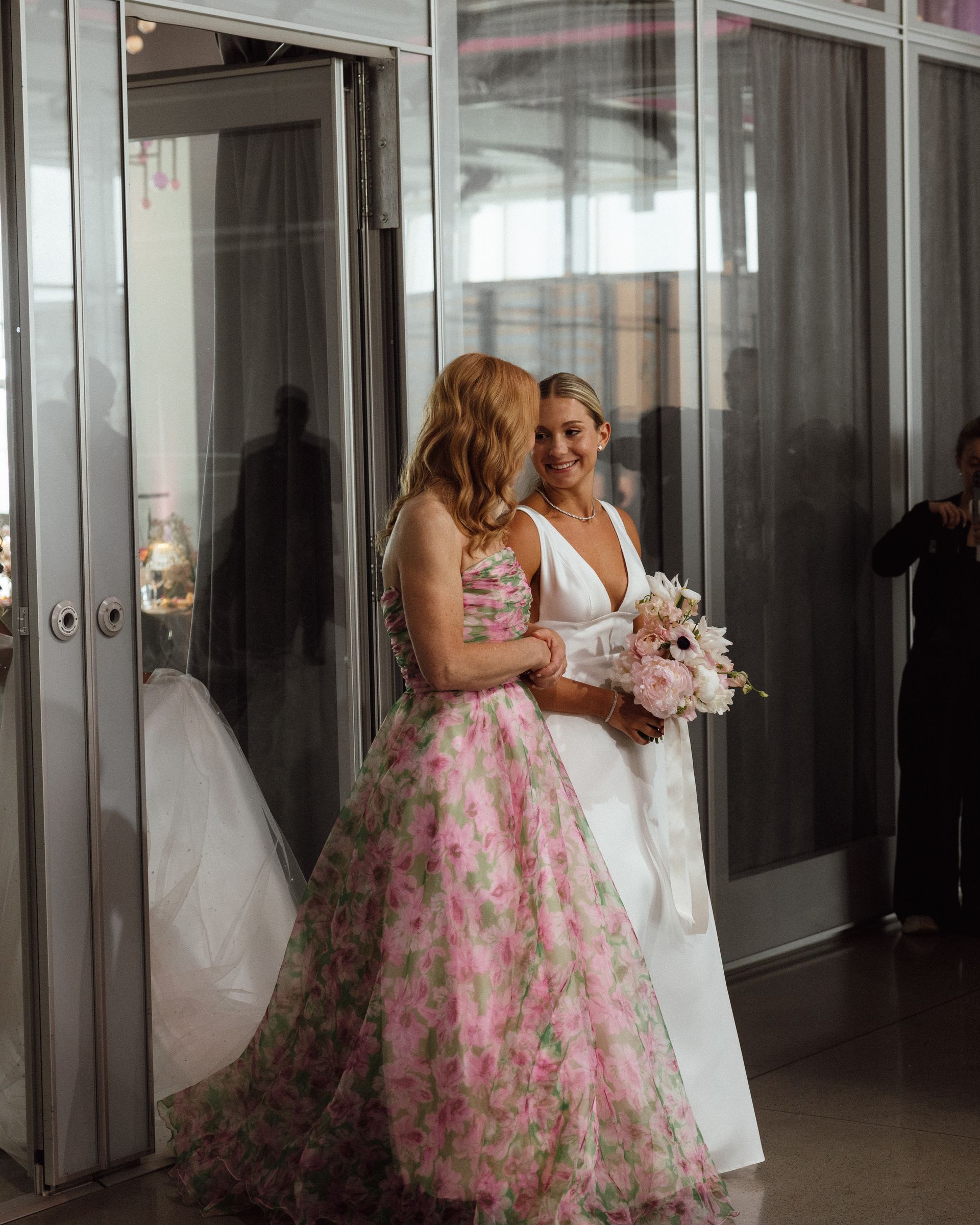 Two women, one in a floral gown, the other in white, embrace near a glass door.