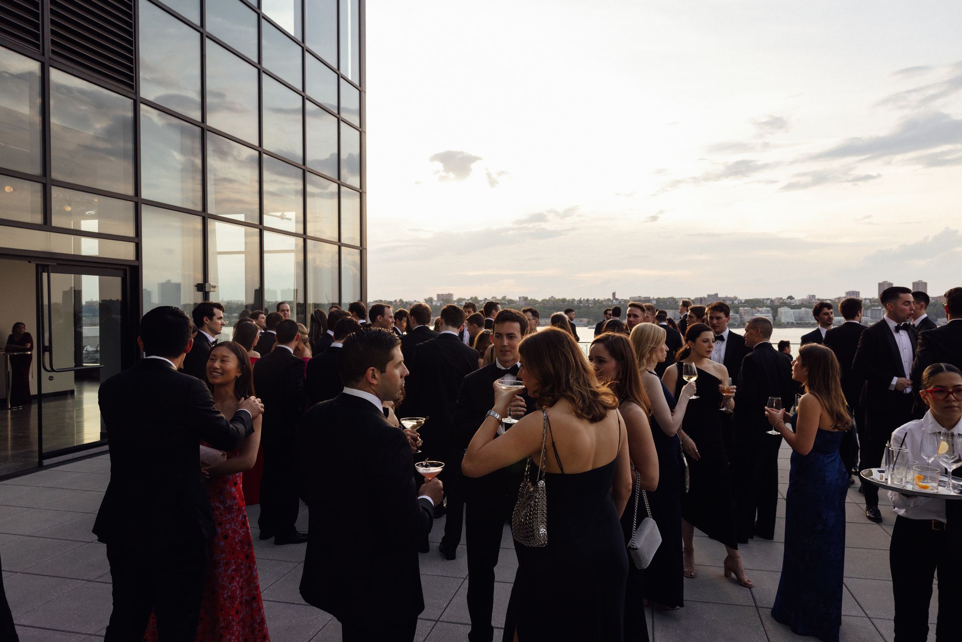 People in formal attire on a rooftop at dusk, socializing. Drinks are being served, with a city view.