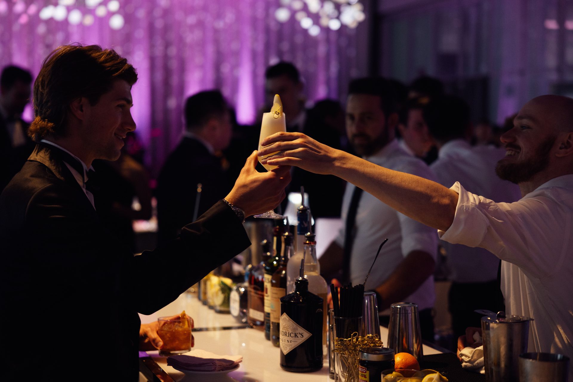 Man in suit receives cocktail from bartender at a well-lit bar; bottles and glasses on display.