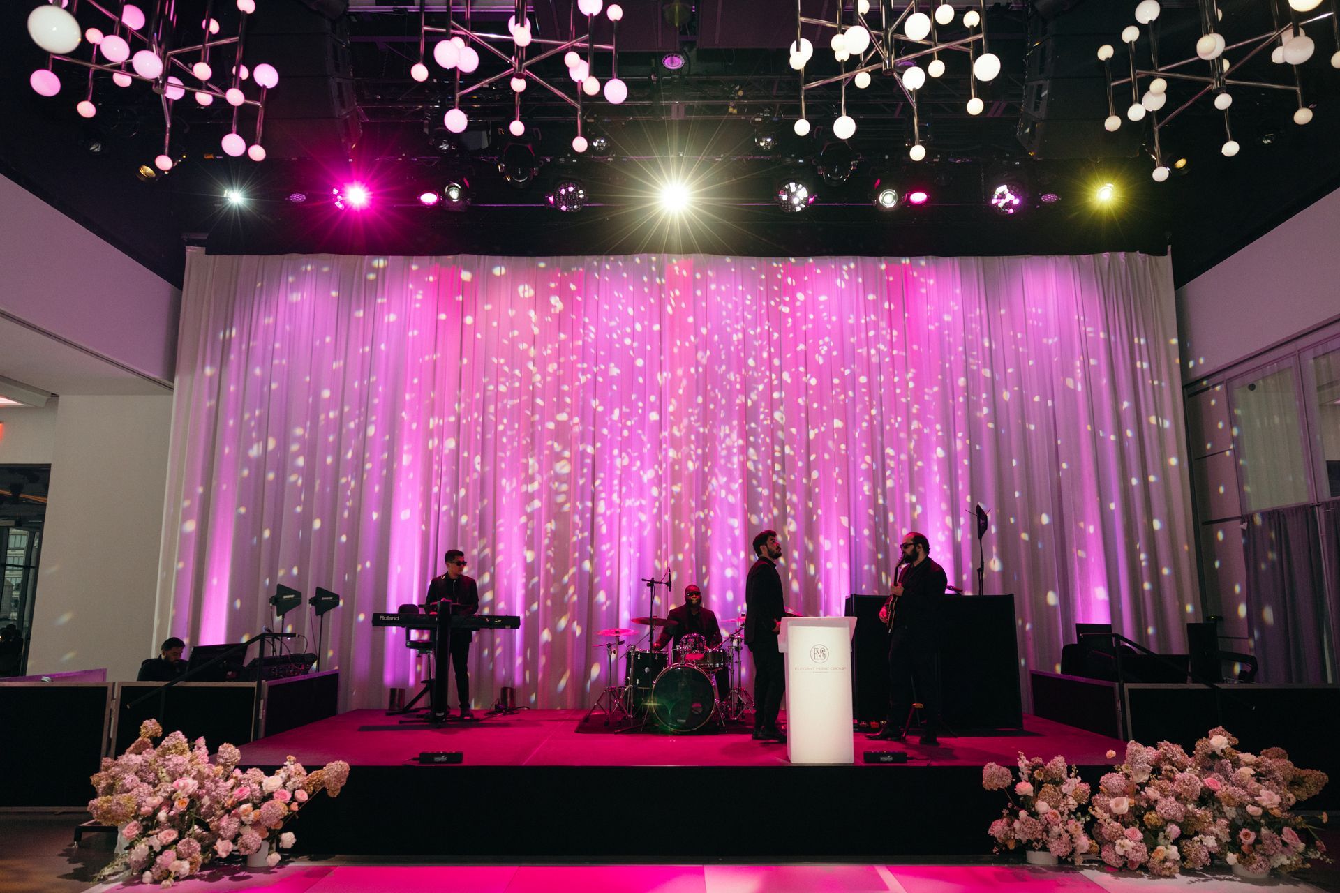 Band performing on stage with pink lighting and curtain backdrop.