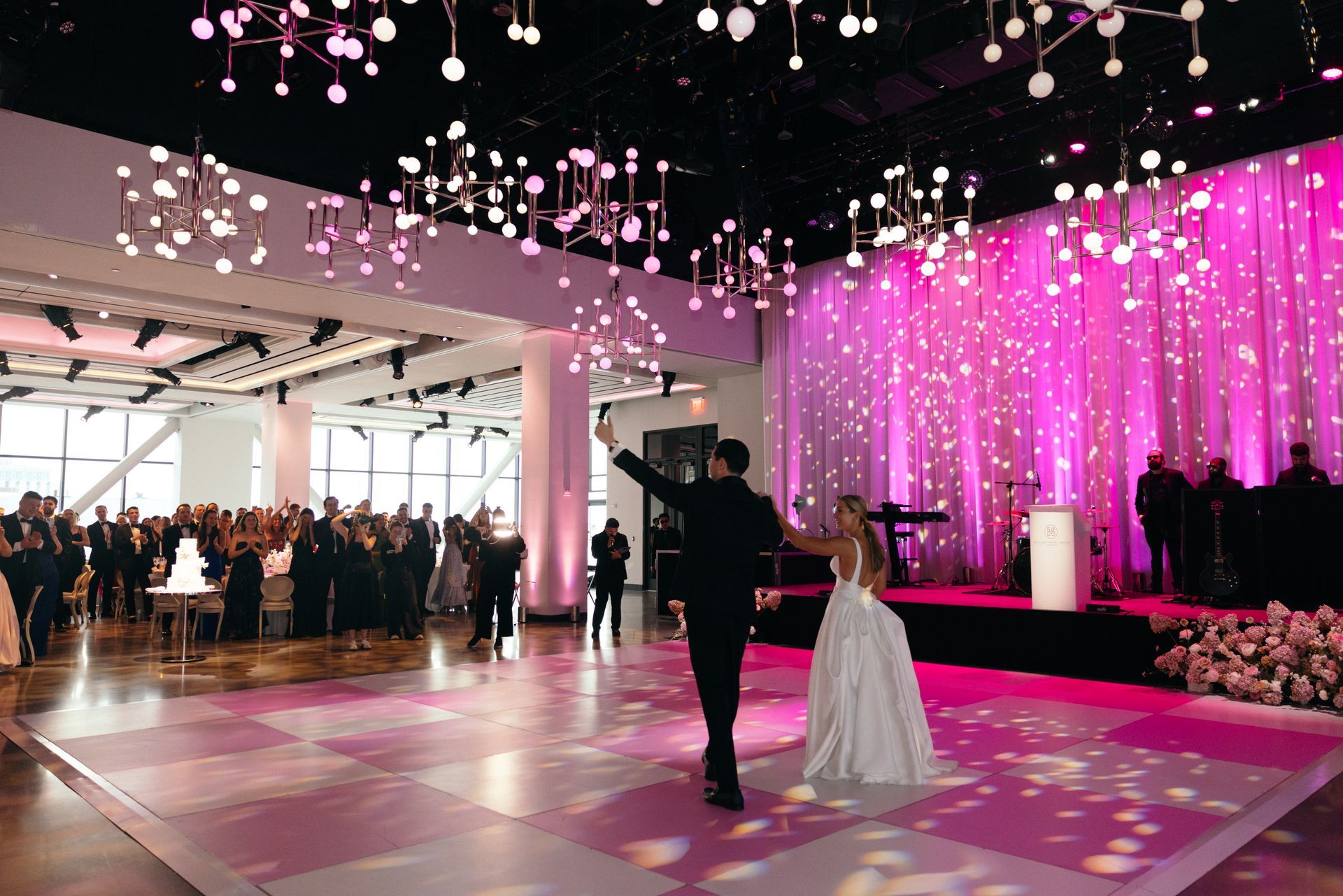 Couple dances at a wedding reception on a pink checkered dance floor, under pink lighting and hanging lights.