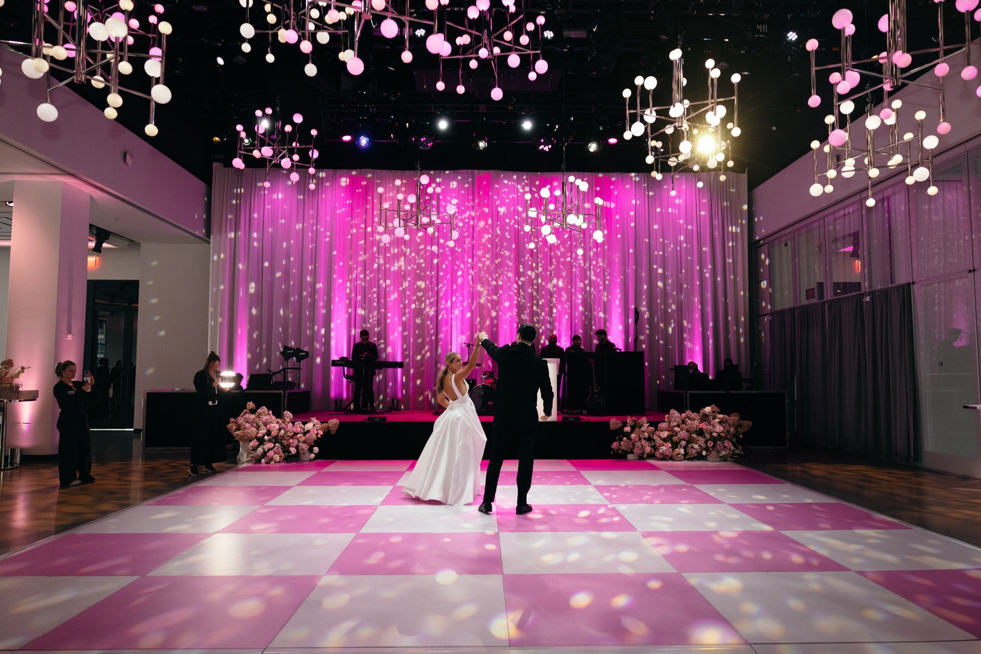 Couple dancing on checkered dance floor with pink backdrop and decorative lights.