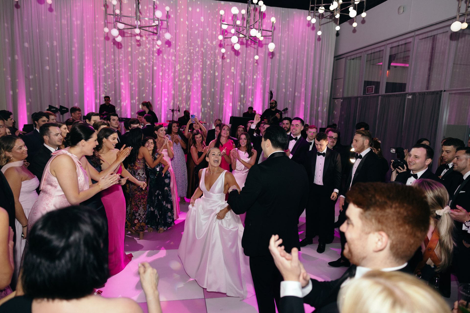 Wedding reception with bride, groom, and guests dancing on a checkered dance floor, illuminated with pink lighting.