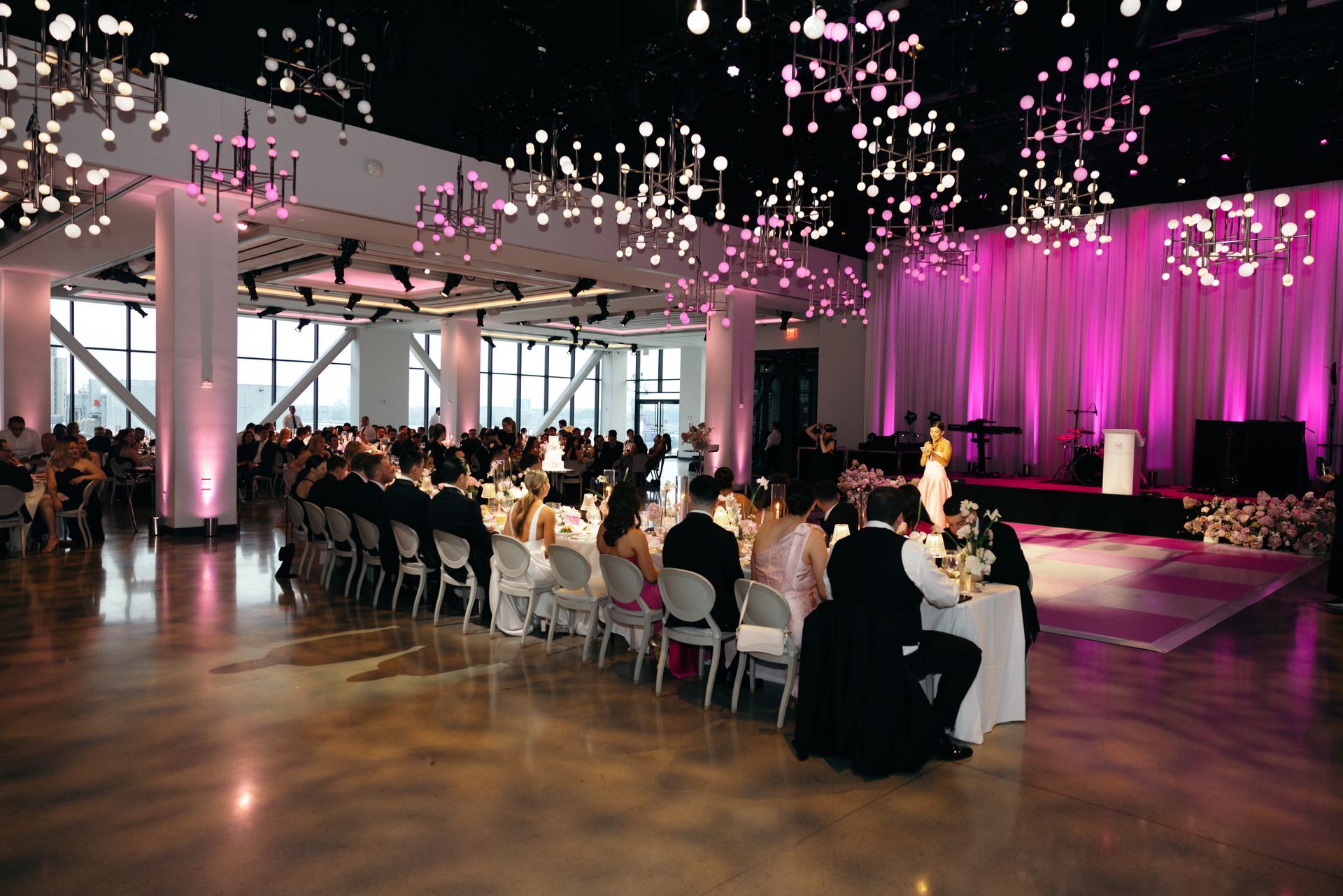 Elegant event space with a long table, guests, stage, and pink lighting.