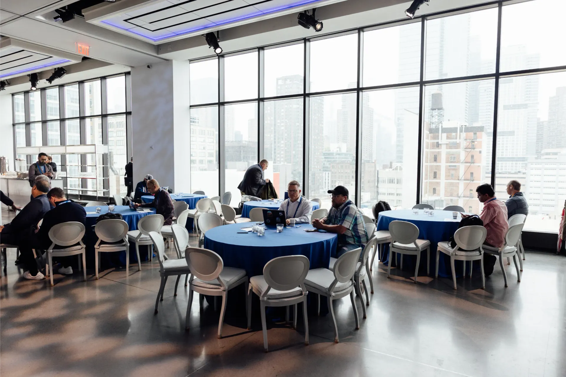 People seated at round tables in a room with large windows, overlooking a cityscape. Blue tablecloths, white chairs.