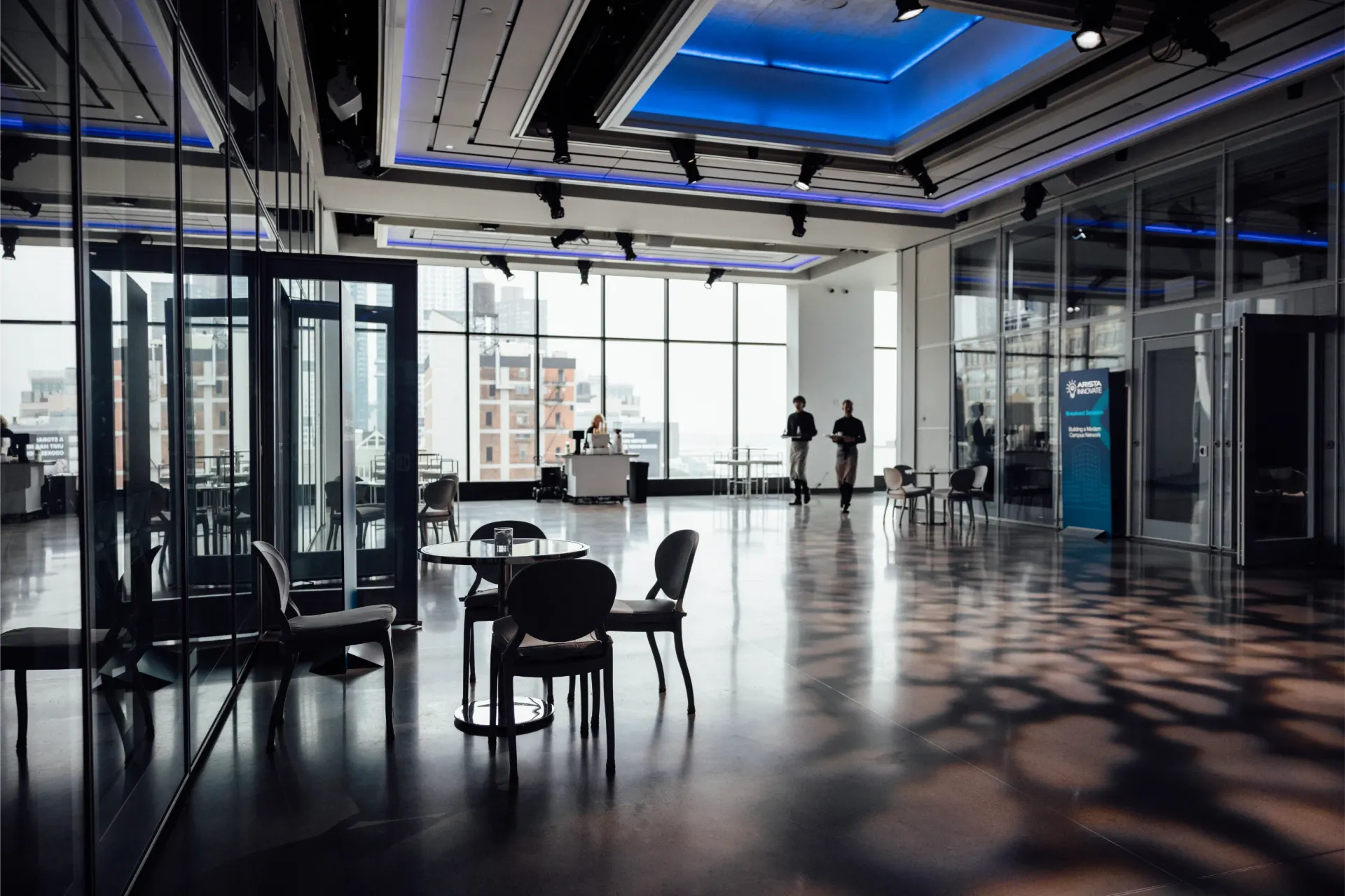 Modern, spacious room with floor-to-ceiling windows. Tables, chairs, and reflective surfaces. Blue ceiling lighting.