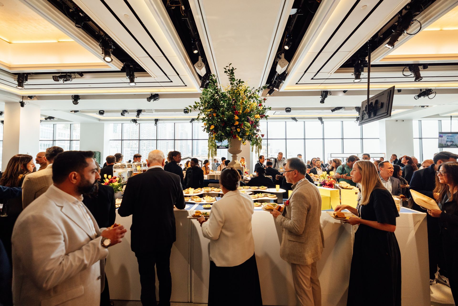 People at a reception, standing and mingling. Tables with food and a floral centerpiece. Brightly lit venue with large windows.