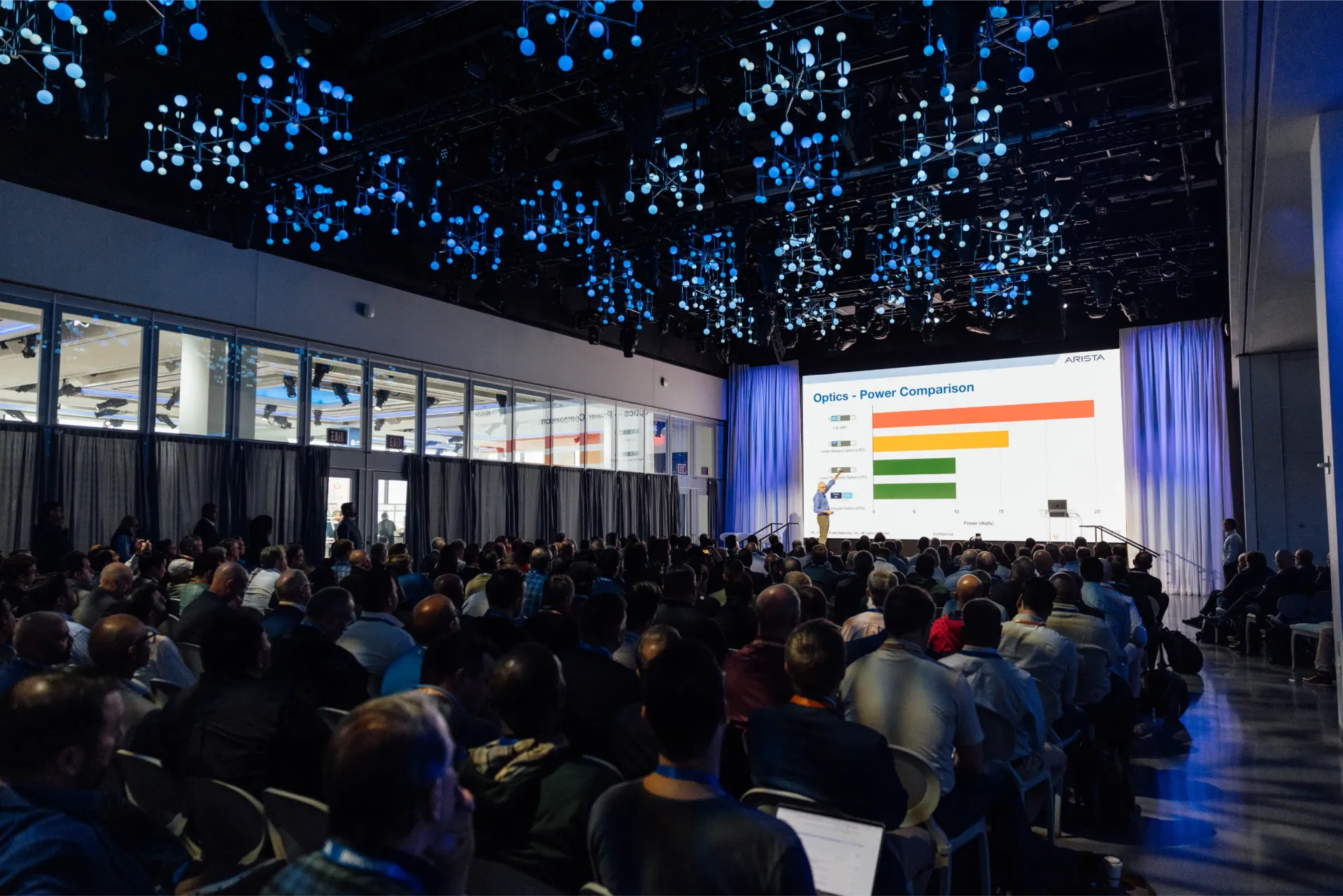 Audience in conference hall; presenter on stage shows data graph; blue overhead lights.
