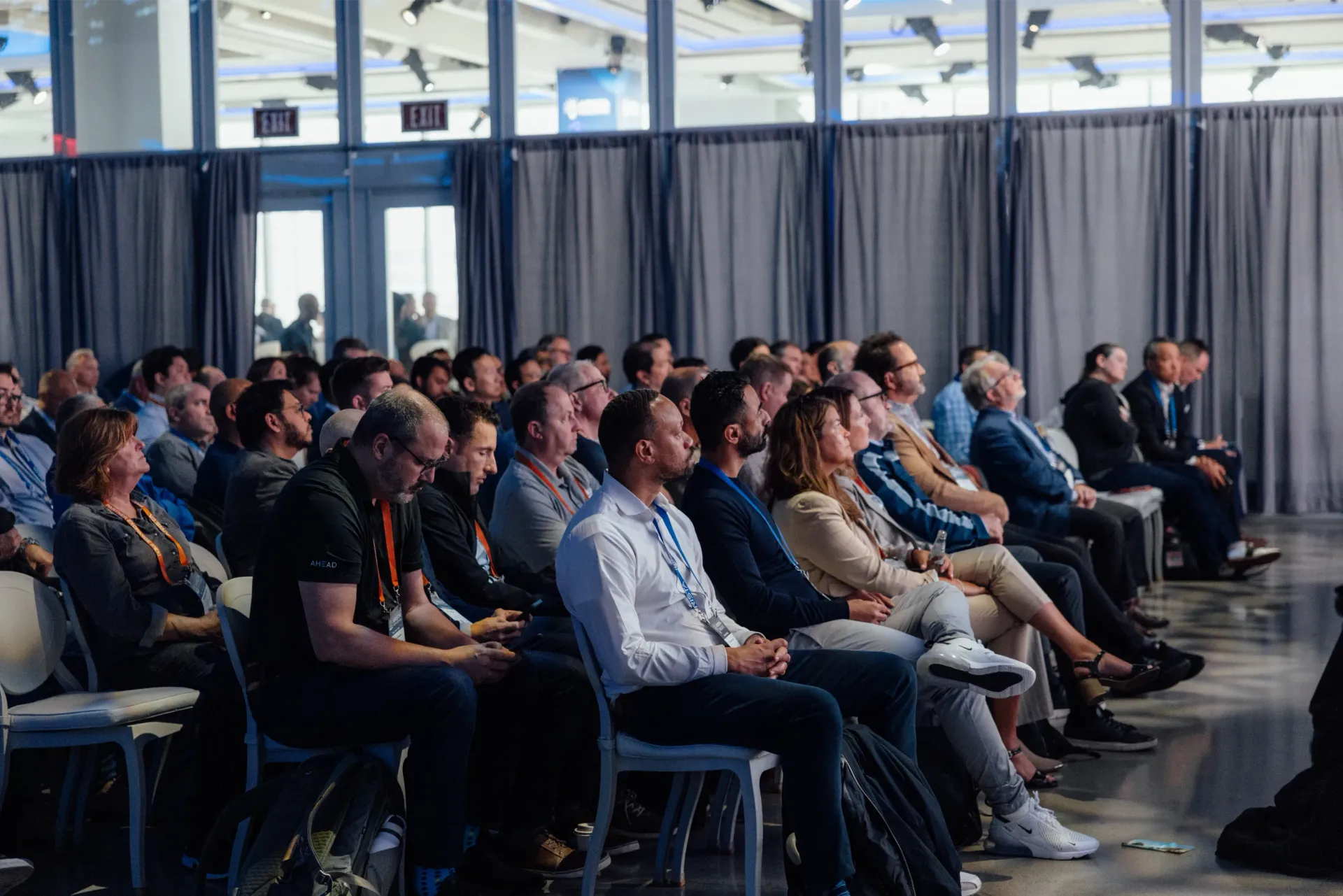 Audience in a conference room listening to a presentation. People are seated and looking forward.