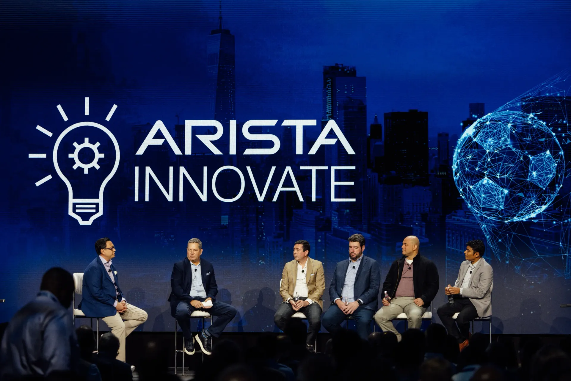Panel at Arista Innovate conference, blue backdrop with logo, speakers seated, discussing.