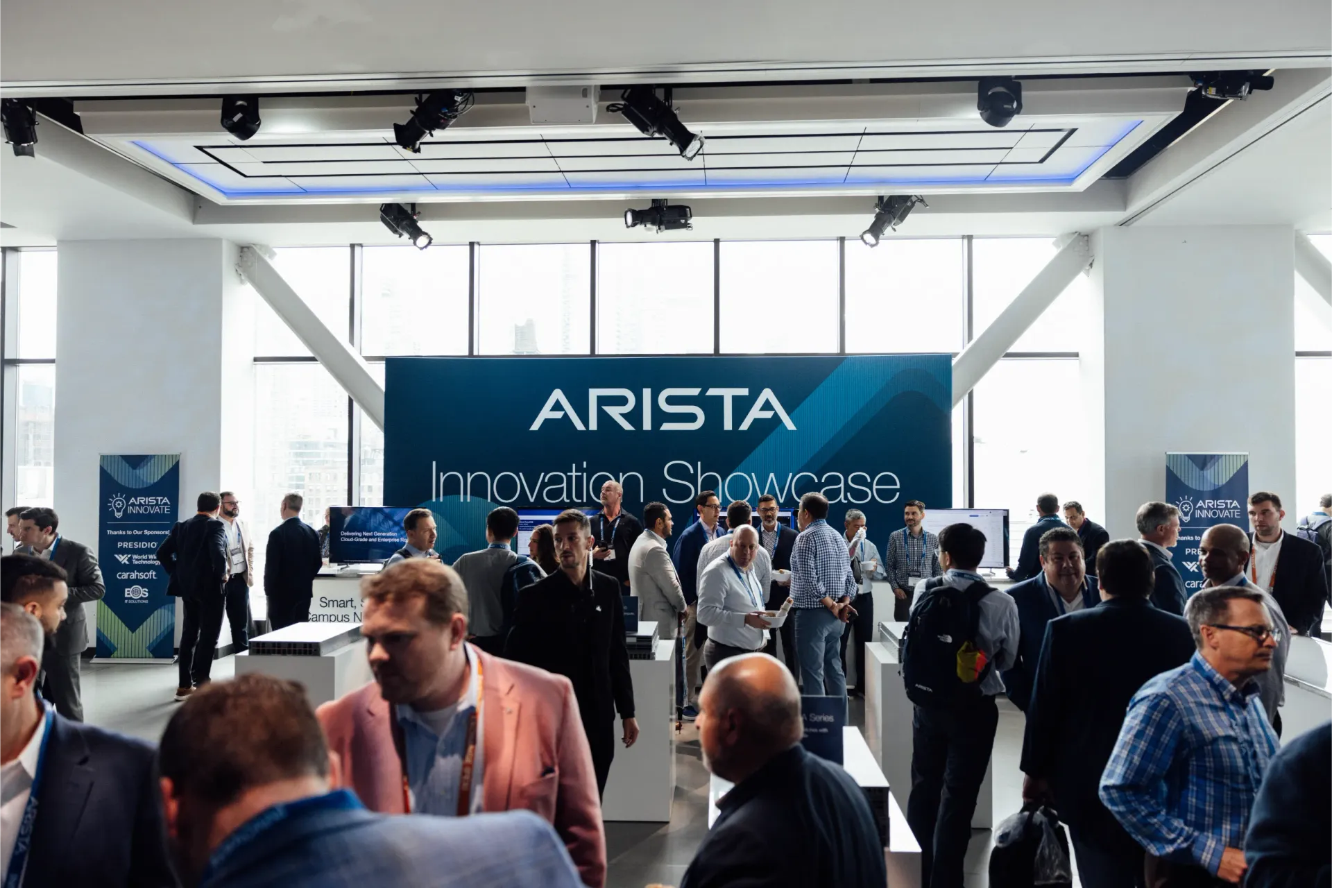 Crowd at Arista Innovator Showcase with banner and display screens in a bright room.