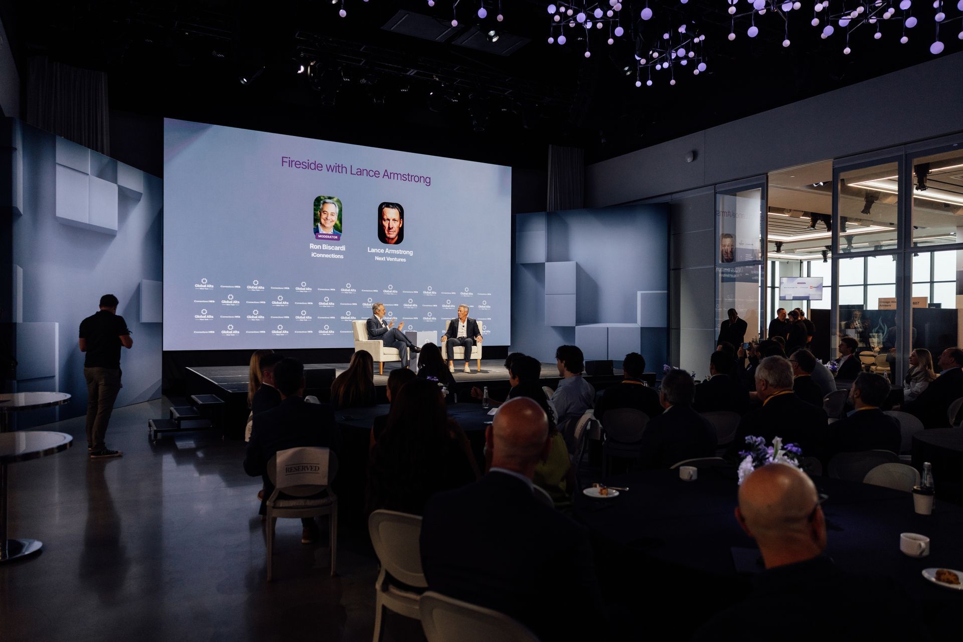Panel discussion on stage in a conference hall, with an audience watching. Screen in background displays speaker names.