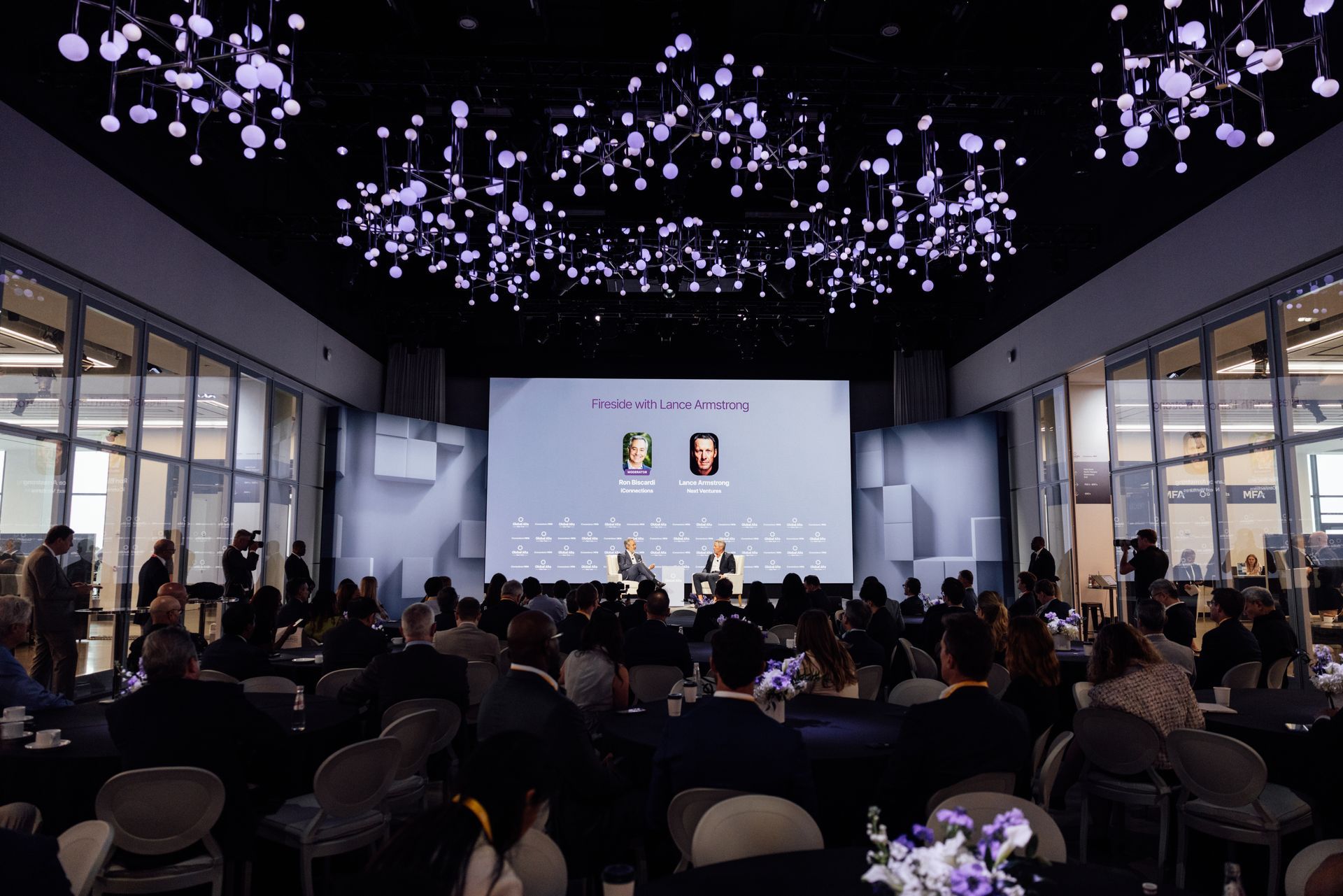 Conference in a modern venue with speakers on stage, audience seated. Large screen displays logos.
