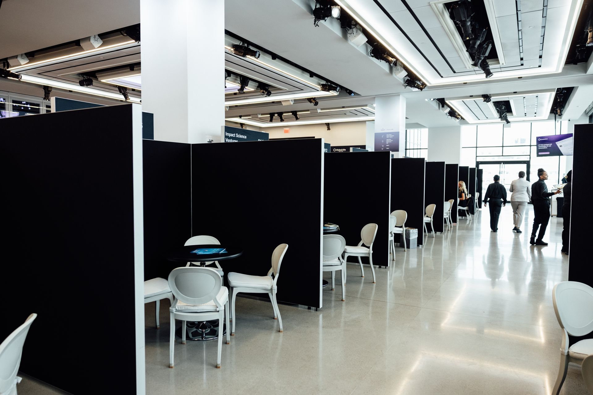 An interior hallway with black dividers creating small spaces, white chairs, and people walking.