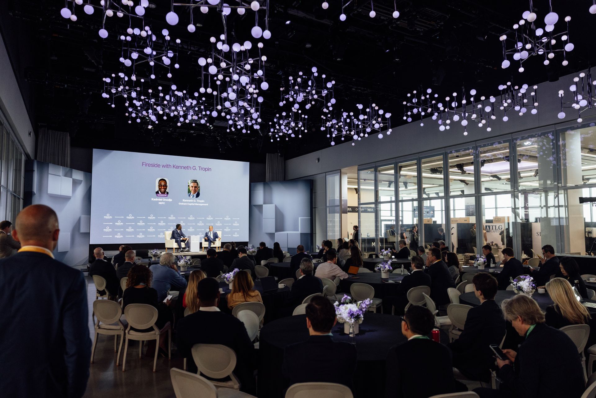 Audience at a conference with speakers on a stage. Decorative lights hang from the ceiling.