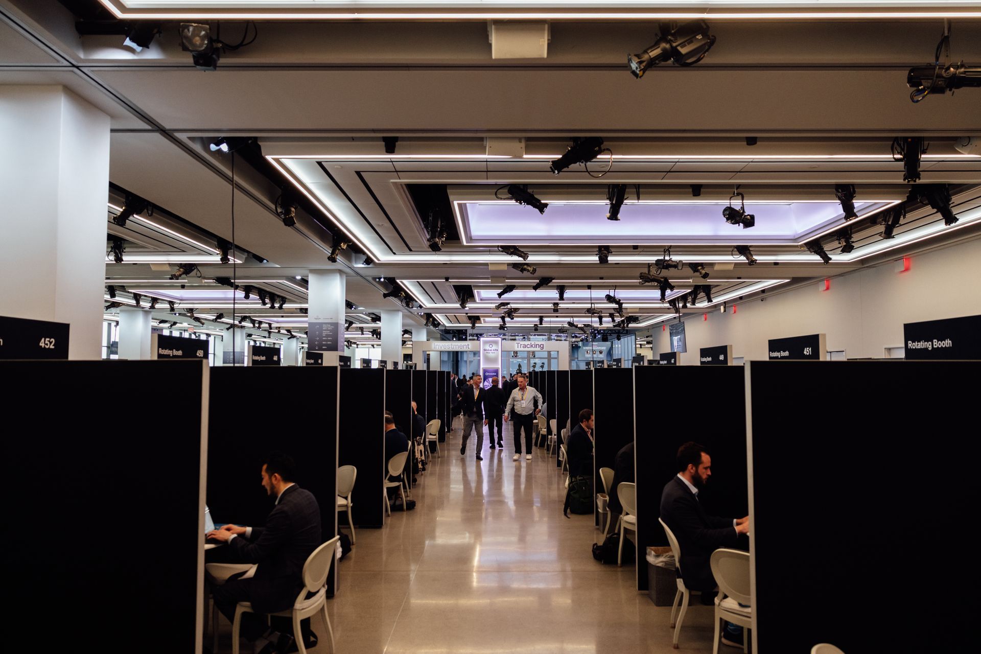 Rows of people working at desks in a large, bright room. Black dividers separate workspaces.