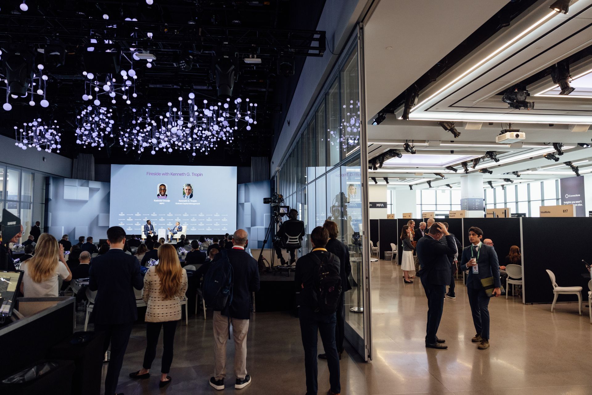 Conference room with a panel on stage, audience, and attendees near a glass wall.