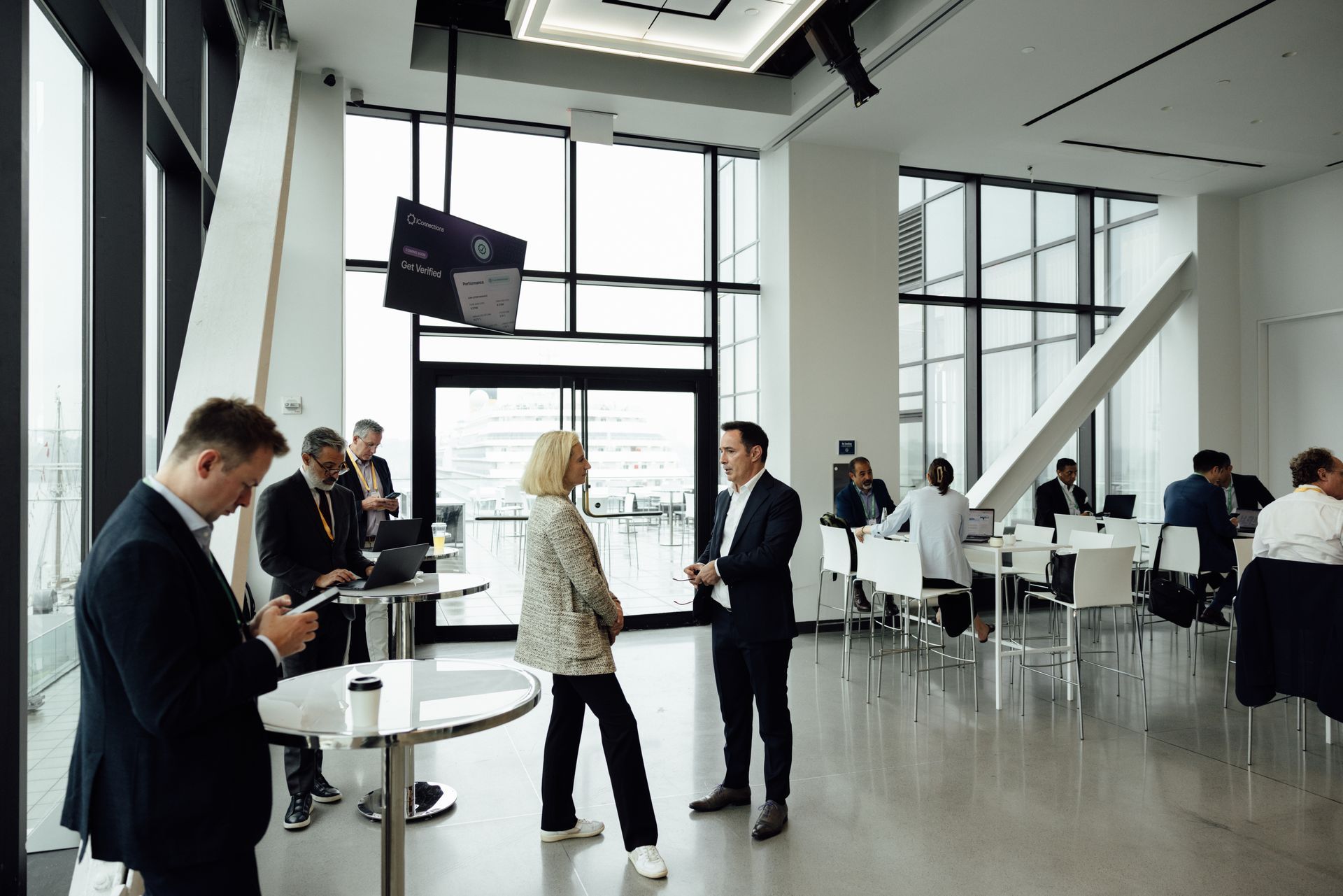 People networking in a bright, modern room with large windows. Several tables and chairs are visible.